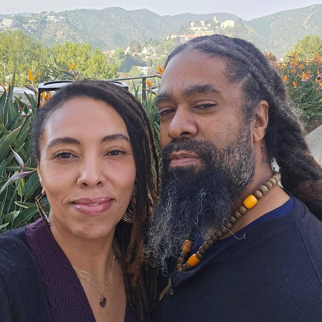 Shylah Hamilton-Touré and Hodari Touré smiling together outdoors in a garden, with green hills behind them.